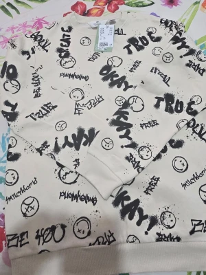 Beige sweatshirt med graffiti-print H&M - Cool beige sweatshirt från H&M med svart graffiti-inspirerat print och smileys över hela tröjan. Rund hals, ribbade muddar och loose passform. Perfekt för dig som gillar streetstyle och vill sticka ut med ett unikt mönster.