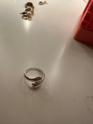 Silvrig ring med böjd design - Säljer en stilren silvrig ring med unik böjd form. Ringen har en modern och minimalistisk look som passar perfekt för dig som gillar enkla men snygga accessoarer. Materialet är metall med blank finish och den är justerbar i storlek.