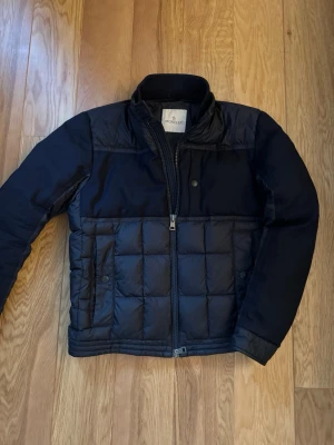 Svart pufferjacka från Moncler - Snygg svart pufferjacka från Moncler med quiltad design på nedre delen och slät ovandel. Jackan har hög krage, dragkedja framtill och tryckknappar vid fickorna. Materialet är polyester och jackan har en clean, modern look som passar perfekt till kyliga dagar.