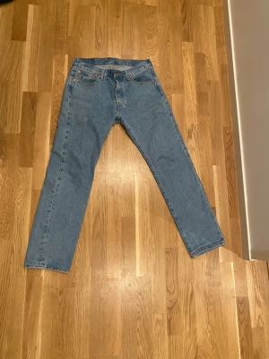 Levi's 501 blå jeans W29 L30 - Klassiska Levi's 501 jeans i ljusblå denim med raka ben och normal passform. Modellen har fem fickor, knappgylf och den ikoniska läderpatchen bak i midjan. Perfekt för dig som gillar tidlös stil och vill ha ett par schyssta jeans i bomull.