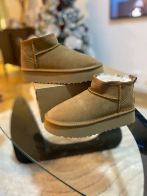 Beige UGG boots med platt sula - Säljer ett par beige UGG boots med platt sula och rund tå. Skorna är tillverkade i mjuk mocka och har vitt fluffigt foder på insidan för extra värme. Klassisk UGG-logga baktill och grov yttersula som ger bra grepp. Perfekta för kalla dagar.