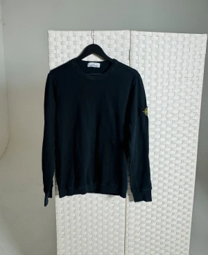 Svart sweatshirt från Stone Island - Svart långärmad sweatshirt från Stone Island med klassisk rund hals och ikonisk patch på vänster ärm. Tröjan är i mjuk bomull och har en normal passform. Perfekt för dig som gillar stilrena och enkla plagg med tydlig streetkänsla.