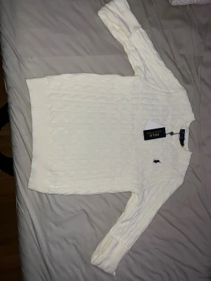 Vit kabelstickad tröja Polo Ralph Lauren - Kabelstickad vit tröja från Polo Ralph Lauren med klassisk rund halsringning och broderad logga på bröstet. Långärmad modell i mjuk bomull som passar perfekt till jeans eller kjol. Stilren och tidlös design som alltid funkar.