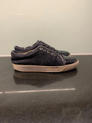 Svarta sneakers från Lanvin - Lanvin sneakers i svart mocka med blankt skinn på tån och vita gummisulor. Klassisk rund tå och snörning med metallöglor. Snygg kontrast mellan material och färg, perfekt för dig som gillar stilrena och lyxiga sneakers. Storlek UK 8 = 43-44