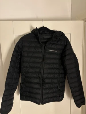 Svart pufferjacka Peak Performance - Svart pufferjacka från Peak Performance med huva och dragkedja framtill. Jackan har ett quiltat mönster och är tillverkad i syntetmaterial. Perfekt för kyliga dagar och har en stilren look med diskret logga på bröstet.