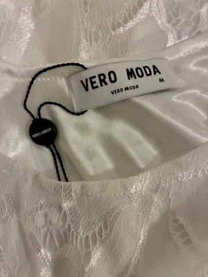 Vit spetsklänning från Vero Moda - vit klänning från Vero Moda i storlek M. Klänningen har ett fint blommigt spetsmönster och rundad halsringning. Den är kort i modellen och har ett lätt, flowigt tyg med fodrad insida som ger en fräsch och somrig känsla. Reflekterar bra i ljus.