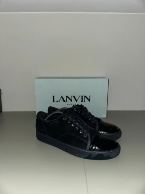 Svarta sneakers från Lanvin med lackad tå - Säljer ett par svarta Lanvin sneakers i mocka och kalvskinn med snygg lackad tå. Skorna har svarta snören, metallöljetter och en platt sula i gummi. Perfekta för dig som gillar stilrena och lyxiga detaljer. Box medföljer och 2 dust bags samm nya snören! Samt kvitto från Care of Carl