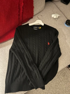 Svart kabelstickad tröja Polo Ralph Lauren - Svart kabelstickad tröja från Polo Ralph Lauren i storlek M. Klassisk design med rund hals och rött broderat Polo-logo på bröstet. Tröjan har långa ärmar och ribbade muddar vid ärmslut och nederkant. Perfekt för dig som gillar stilrena och tidlösa plagg.