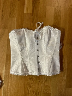 Vit korsett med spets och snörning - Säljer en vit korsett med vackert broderat mönster och glansig yta. Toppen har snörning i ryggen, metallknappar framtill och volangkant upptill och nedtill. Perfekt för dig som vill ha en statement piece med romantiska detaljer och snygg passform. Storlek XL men skulle säga att den är liten i storleken då jag brukar ha M i tröjor 