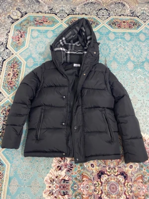 Svart pufferjacka från Burberry - Svart pufferjacka från Burberry med avtagbara ärmar så du kan bära den som väst. Jackan har huva med grått och vitt rutigt foder, dragkedja och knappar framtill samt två sidofickor med dragkedja. Märkespatch på ena ärmen och klassisk Burberry-tag.