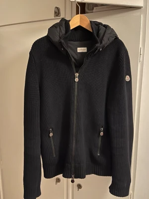 Svart pufferjacka från Moncler - Svart pufferjacka från Moncler med stickade ärmar och quiltad kropp. Jackan har huva, två dragkedjefickor fram och ribbade muddar. Materialmix av dun och stickad polyester ger både värme och stil. Perfekt för kyliga dagar och snygg till streetwear.