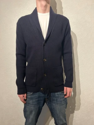 Mörkblå stickad kofta med knappar -  mörkblå stickad kofta med, stora framfickor och fyra knappar framtill. Koftan har en bred sjalkrage och lång ärm, perfekt för lager på lager under kyliga dagar. Tröjan är från pier one och är i så fint skick de går använd en gång. Står inte vilken storlek och kommer it ihåg vilken jag köpte men tror M. är 183