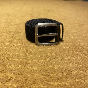 Svart JACK & JONES flätat bälte - Bilderna visar ett svart flätat bälte med silvrigt spänne och detaljer i skinn, inte en väst. Kan ej ge beskrivning eller taxonomier för väst.