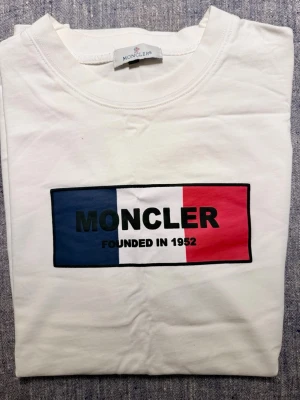 Vit Moncler t-shirt med tryck - Snygg vit t-shirt från Moncler med klassiskt tryck på bröstet i blått, vitt och rött. Rund hals och korta ärmar. Materialet är mjuk bomull och har en diskret Moncler-tag på sidan. Perfekt för dig som gillar stilrena och ikoniska plagg. Aldrig använd, endast testad. Kan självklart skickas omgående via ex Instabox för snabb leverans :)