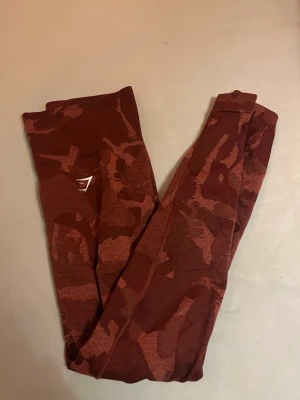 Mörkröda camo leggings från Gymshark - Snygga mörkröda leggings från Gymshark med camouflagemönster och hög midja. Stretchigt syntetmaterial som sitter tight och är perfekt för träning. Diskret logga på höften. Superskön passform och cool design.