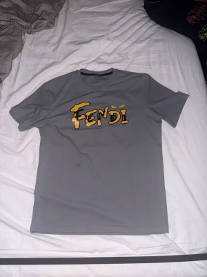 Grå Fendi t-shirt med tryck - Snygg grå t-shirt från Fendi med korta ärmar och rund hals. Framsidan har ett stort Fendi-tryck i gult och svart, och baksidan har Fendi-ögon i gult vid nacken. Materialet känns mjukt och lätt, perfekt för en chill stil.