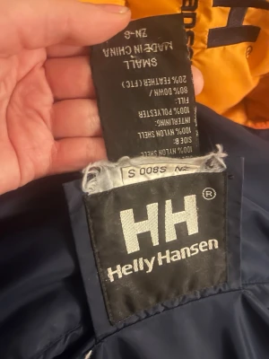 Helly Hansen pufferjacka, blå/orange, S - Säljer en fet pufferjacka från Helly Hansen i storlek S. Jackan är vändbar med ena sidan i mörkblått och andra i orange, båda med HH-logga på bröstet. Dragkedja i orange och snörning nedtill. Fylld med dun och fjäder, perfekt för kalla dagar. 