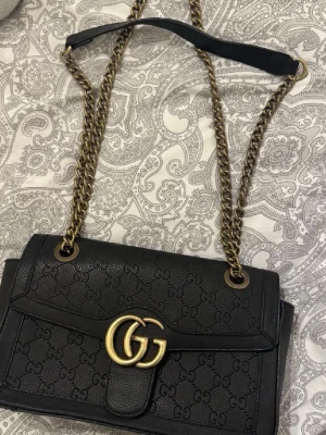 Svart axelväska från Gucci i skinn - Säljer en svart axelväska från Gucci med GG-logga i guld framtill och präglat GG-mönster över hela väskan. Väskan har kedjerem i antikguld och innerfoder i beige. Perfekt storlek för det viktigaste och har dragkedja inuti.
