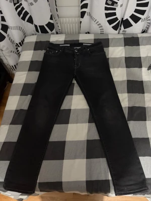 Jacob Cohen svarta jeans style 688 - Jacob Cohen svarta jeans, style 688, med kontrasterande vita sömmar och klassisk femficksdesign. Jeansen har knappgylf och är tillverkade i ett mjukt bomullsmaterial. Passformen är rak och de har en stilren look med diskret logopatch bak på midjan. Dock står det 35 men sitter 33/32 eftersom jag är 185 78 o sitter prima