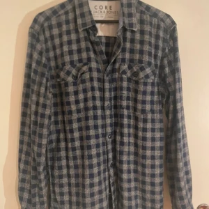 Rutig blå skjorta från Jack & Jones - Säljer en rutig skjorta från Jack & Jones Core med blå och grå toner. Skjortan har långa ärmar, klassisk krage och två bröstfickor med knappar. Materialet är mjukt och känns som bomull, perfekt för dig som gillar avslappnad stil.