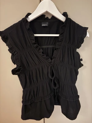 Svart blus med volanger från Gina Tricot - Säljer en svart blus från Gina Tricot i storlek S. Blusen har volangdetaljer längs ärmar och hals, omlott-design med knyt framtill och är tillverkad i ett lätt, strukturerat tyg. Perfekt för dig som gillar romantiska detaljer och en feminin look.