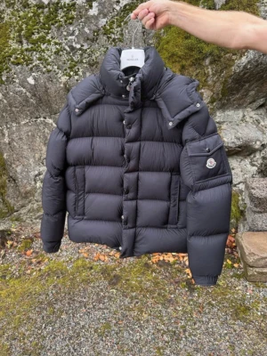 Moncler vezere - Säljer en svart pufferjacka från Moncler med hög krage och avtagbar huva. Jackan har långa ärmar, dragkedja och knappar framtill samt en ficka med logotyp på ärmen. Perfekt för kalla dagar och riktigt snygg streetstil.