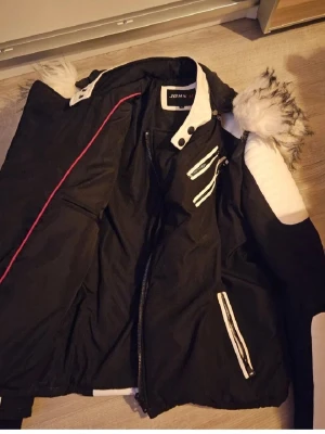 Svart och vit pufferjacka från John H - Snygg svart och vit pufferjacka från John H med vita detaljer och dragkedjor. Jackan har fluffig fuskpäls på huvan, quiltade vita partier på axlarna och flera fickor med dragkedja. Perfekt för dig som vill sticka ut med en cool vinterstil.