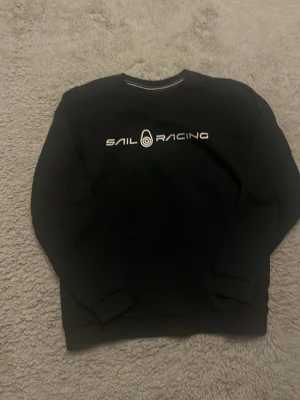 Svart Sail Racing sweatshirt - Svart sweatshirt från Sail Racing med vit logotyp tryckt på bröstet och en mindre logga i nacken. Tröjan har rund hals, långa ärmar och ribbade muddar vid ärmslut och nederkant. Perfekt för en chill och sportig stil.
