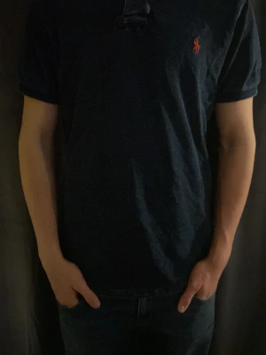 Mörkblå kortärmad piké från Polo Ralph Lauren - Säljer en mörkblå piké från Polo Ralph Lauren med klassisk passform och korta ärmar. Pikén har den ikoniska röda loggan broderad på bröstet och är gjord i mjuk bomull. Perfekt till jeans eller shorts för en clean och enkel look.