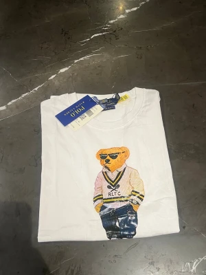 Vit Polo Bear t-shirt från Ralph Lauren - Vit t-shirt från Ralph Lauren med ikoniskt Polo Bear-tryck framtill. Klassisk rund hals och korta ärmar. Skön bomullskvalitet och avslappnad passform. Perfekt för dig som gillar streetstyle och vill sticka ut med en cool printad björn.