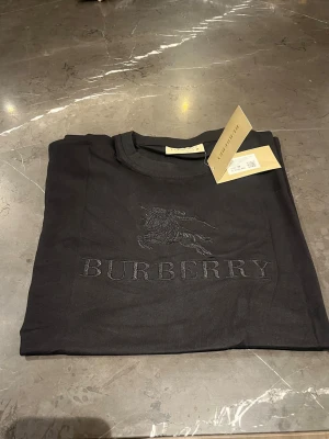 Svart Burberry t-shirt med broderi - Säljer en svart t-shirt från Burberry i mjuk bomull. T-shirten har korta ärmar och ett snyggt broderat Burberry-logo och motiv på bröstet. Klassisk rund hals och stilren design, perfekt för dig som gillar exklusiva märken.
