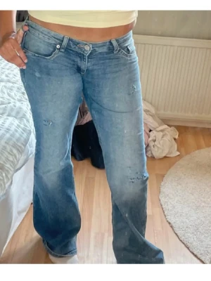 Lågmidjade jeans - Säljer ett par blå bootcut jeans med lätt slitna detaljer och klassisk femficksdesign. Jeansen har låg midja och är tillverkade i mjukt denimmaterial som ger en skön känsla. Perfekt för dig som gillar en avslappnad och trendig stil.