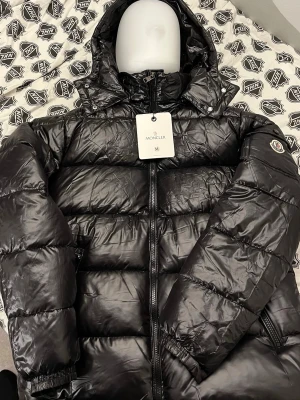 Moncler Maya  - Svart pufferjacka från Moncler i blankt syntetmaterial med huva och dragkedja. Jackan har Moncler-logga på ärmen och coolt serietidningsmotiv på insidan. Perfekt för dig som vill ha en varm och snygg jacka med streetkänsla.