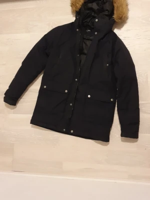 Svart parkas från Everest med fuskpäls - Säljer en svart parkas från Everest i storlek XXS. Jackan har en mysig huva med fuskpäls, stora fickor med knappar och dragkedja framtill. Insidan är quiltad och fodrad för extra värme. Perfekt för kalla dagar när du vill hålla stilen.