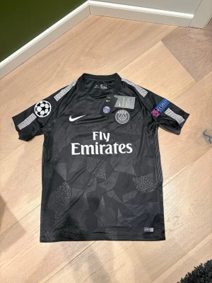 Psg borta tröja 2017 - Bilderna visar en svart PSG fotbollströja med Neymar Jr och nummer 10, samt Champions League och Respect-emblem. Ingen sko finns med på bilderna.