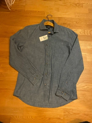 Blå skjorta från Hampton Republic - Snygg blå skjorta från Hampton Republic. Helt ny med prislapp (Orginal pris 499kr) i storlek S, regular fit med klassisk krage och bröstficka. Skjortan har långa ärmar och knappar framtill samt vid manschetterna. Perfekt för dig som gillar stilrena plagg.