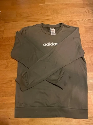 Olivgrön sweatshirt från Adidas - Säljer en olivgrön sweatshirt från Adidas i storlek S i utmärkt skick. Tröjan har klassisk rund hals, långa ärmar och vit/silvrig Adidas-logga tryckt på bröstet. Perfekt för chill eller sportiga dagar.