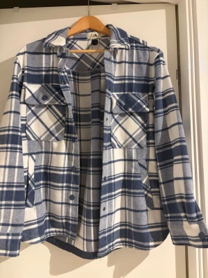 Blåvit rutig overshirt från C&A - Säljer en blåvit rutig overshirt från C&A med klassisk krage, två stora bröstfickor med knappar och långa ärmar. Jackan har en loose passform och är gjord i mjuk bomull, perfekt att slänga över en hoodie eller t-shirt. Stängs med tryckknappar framtill.