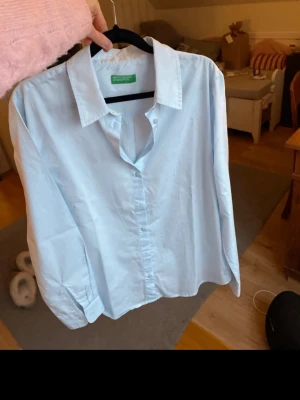 Ljusblå skjorta från Benetton - Snygg ljusblå skjorta från United Colors of Benetton med klassisk krage och knappar framtill. Skjortan har lång ärm och är gjord i mjuk bomull. Helt oanvänd