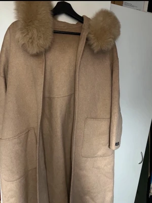 Beige kappa med päls och bälte - Säljer en beige kappa med fluffig pälsdetalj på huvan och stora fickor framtill. Kappan har ett bälte i midjan för snygg passform och är tillverkad i mjukt ullmaterial. 
