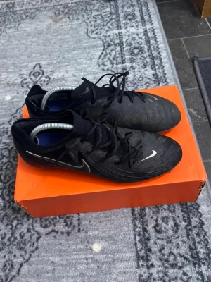 Phantom Gx Pro FG - Säljer ett par svarta Nike fotbollsskor med snyggt mönster och vita detaljer på sidan. Skorna har snörning och en strumpliknande krage för extra komfort. Ovandelen är tillverkad i syntetmaterial och har ACC-teknologi för bra grepp i alla väder. Perfekt för dig som vill ha stil på planen.