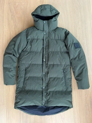 Grön lång pufferjacka med huva - Säljer en mörkgrön pufferjacka med huva och ribbade muddar. Jackan har dragkedja och knappar framtill, två sidofickor med dragkedja och ett stilrent märke på ärmen. Perfekt för kalla dagar och riktigt snygg längd.