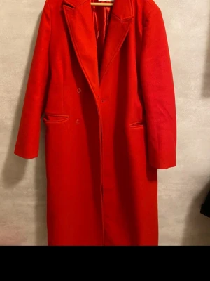 Röd lång kappa från NLY Trend - Säljer en riktigt snygg röd kappa från NLY Trend. Den är lång, har klassisk krage och knappar framtill. Fodrad insida och två fickor på sidorna. Perfekt för dig som vill sticka ut i höst och vinter. Passar till allt och ger en clean look.