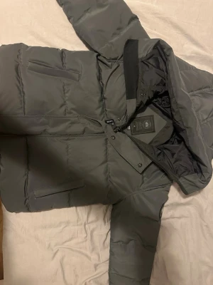 Mörkgrå pufferjacka från Canada Goose McMillan  - Säljer en mörkgrå pufferjacka från Canada Goose med huva och klassisk logga på ärmen. Jackan har dragkedja och knappar framtill, två sidofickor och quiltad design. Perfekt för kalla dagar och riktigt snygg till streetwear-stilen.