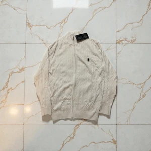 Vit full zip kofta från Polo Ralph Lauren - Säljer en beige kofta från Polo Ralph Lauren med dragkedja framtill och det klassiska broderade logot på bröstet. Tröjan har ett snyggt kabelstickat mönster, ribbade muddar och hög krage. Perfekt för dig som gillar stilren och tidlös design.