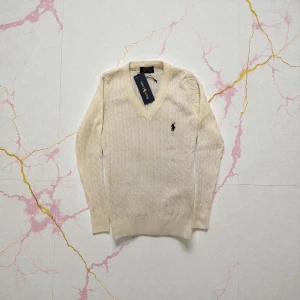 Vringad Beige stickad tröja från Polo Ralph Lauren - Snygg beige stickad tröja från Polo Ralph Lauren med v-ringad hals och kabelstickat mönster. Tröjan har lång ärm och en liten broderad logga på bröstet. Perfekt för dig som gillar klassisk och stilren design.