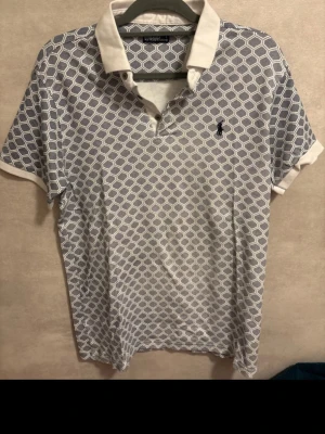 Blåvit polotopp från Ralph Lauren - Säljer en snygg polotopp från Ralph Lauren i blått och vitt med geometriskt mönster. Toppen har klassisk krage, korta ärmar och knappar vid halsen. Märkets ikoniska logga broderad på bröstet. Perfekt för dig som gillar stilrena detaljer.