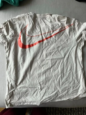 Over size Vit Nike t-shirt med röd swoosh junior - Vit t-shirt från Nike med stor röd swoosh-logga på ryggen och liten grå swoosh på bröstet. Klassisk rund hals och korta ärmar. Tillverkad i mjuk bomull som är skön mot huden. Perfekt för dig som gillar sportig och enkel stil. Juniorstorlek XL 156-168 cm