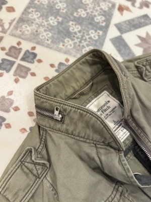 Vintage parkajacka Abercrombie & Fitch - Snygg militärgrön parkajacka från Abercrombie & Fitch med ståkrage och coola detaljer som dragkedja och knappar. Fyra fickor framtill och schyssta sömmar ger jackan en urban vibe. Perfekt för dig som gillar streetstyle och vill ha en jacka med attityd. Storlek L men sitter som s/m.