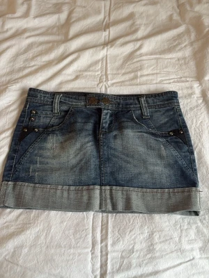 Jeanskjol från Stradivarius, stl 36 - Kort jeanskjol från Stradivarius med slitningar och dekorativa nitar på fickorna. Kjolen har dubbla knappar i midjan, bälteshällor och snygga bakfickor med bruna sömmar och nitar💕